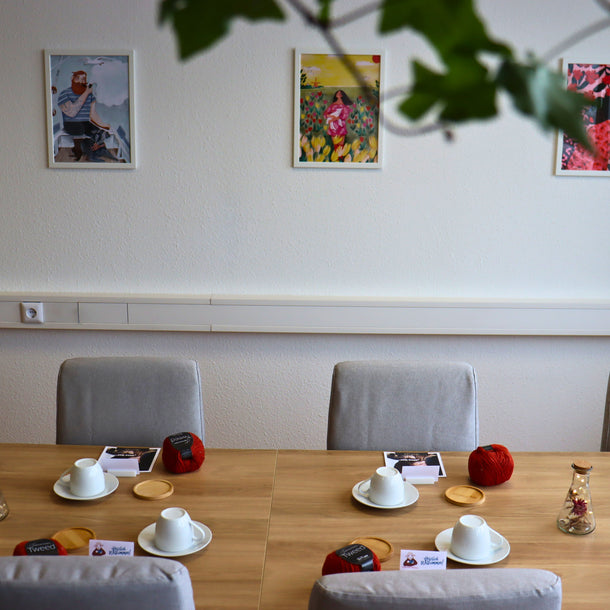 Workshoptisch mit Kaffeetasse, Wolle und Strickanleitungen, an der Wand hängen farbenfrohe Illustrationen
